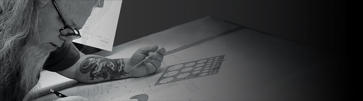 Bearded John Page at drafting table making designs with mechanical pencil, papers, and tools—awesome dragon tattoo visible on his left forearm.